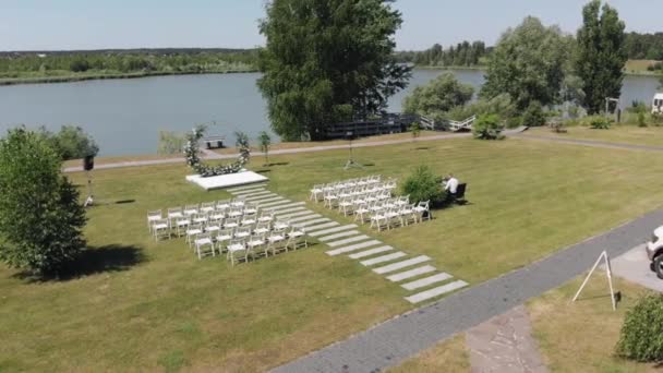 Вид с воздуха на цветочную арку и ряды стульев для свадебной церемонии у реки в летний день — стоковое видео
