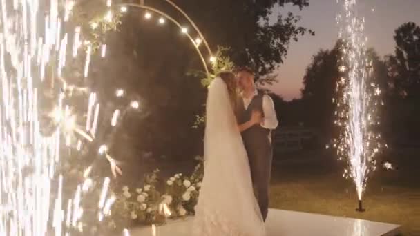A noiva e o noivo dançam perto do arco da flor com lâmpadas acesas e fogos de artifício no chão. Transição para o efeito bokeh. Movimento lento — Vídeo de Stock
