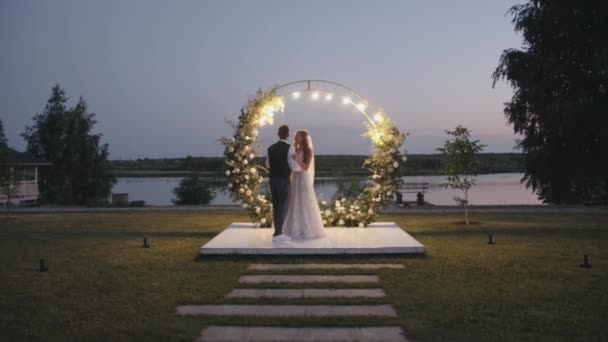 Geëmailleerde bruid en bruidegom dansen op een platform voor een bloemboog versierd met een brandende bloemenslinger op een zomeravond. Langzame beweging — Stockvideo