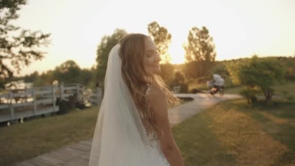 En spektakulär ung brud går till träpiren på flodstranden vid solnedgången och ler över axeln. Långsamma rörelser — Stockvideo