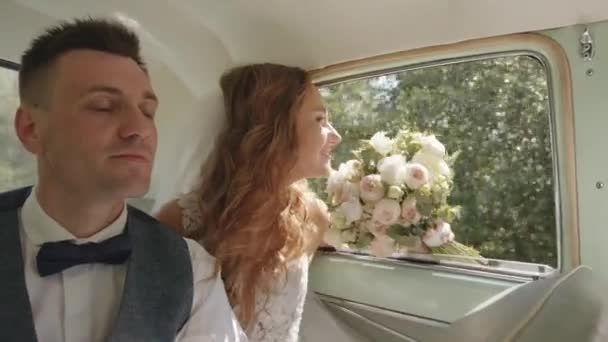 Happy newlywed couple rides in the back seat of a retro car and the bride sniffs a bouquet of flowers. Slow motion — Stockvideo