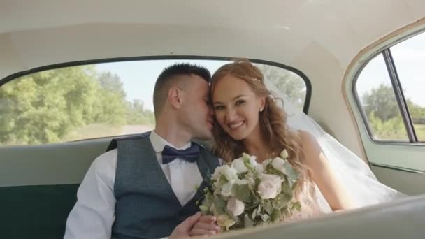 Elegante sposo dice qualcosa nell'orecchio di una sposa affascinante con un bouquet in mano seduto sul sedile posteriore di una macchina retrò. Rallentatore — Video Stock