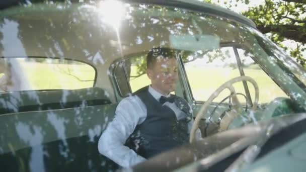 Tânărul om de succes într-o cămașă albă într-o vestă și într-o cravată de arc stă la volanul unei mașini retro în natură. Slow motion — Videoclip de stoc