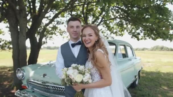Belarus. Gomel region. August - 21, 2021: Happy groom hugs his laughing bride with a bouquet while standing near a retro car under a tree — 图库视频影像