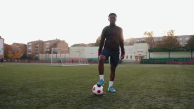 Hoş, genç, siyah bir kız bir stadyumda duruyor ve ayağını bir futbol topunun üzerine koyup güneşin arkasına yaslanıyor. Yavaş çekim