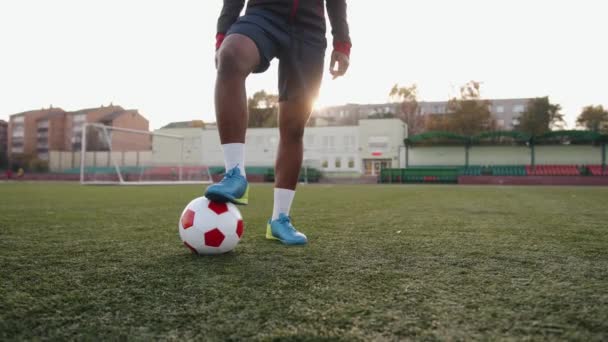 Una piacevole ragazza nera si trova in uno stadio della città e mette il piede su un pallone da calcio sullo sfondo del sole. Rallentatore — Video Stock