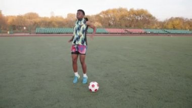 Genç siyah kız futbol sahasında ısınıyor, topun yanında ve güçlü bir şekilde bacaklarını kaldırıyor.