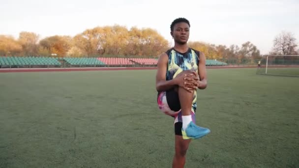 Zelfverzekerde zwarte jonge meisje in helder uniform gaat naar binnen voor sport op het voetbalveld en doet been strekken. Langzame beweging — Stockvideo