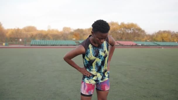 Sonriente joven negra calentándose y haciendo rotaciones e inclinaciones del cuerpo mientras está de pie en el campo de fútbol al lado de la pelota — Vídeos de Stock