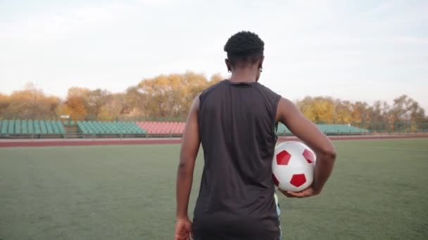 Mladá černoška jde na fotbalové hřiště s míčem v ruce, otočí se ke kameře a usměje se — Stock video