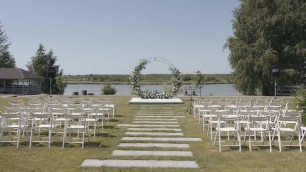 Vista do arco de flores e o caminho entre as fileiras de cadeiras no local da cerimônia de casamento nas margens do rio — Vídeo de Stock