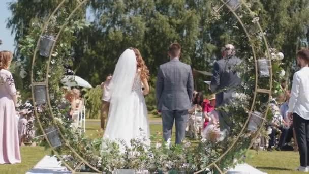 Visão traseira da noiva e do noivo em pé no arco de flores na frente dos convidados e ouvir o discurso na cerimônia de casamento — Vídeo de Stock