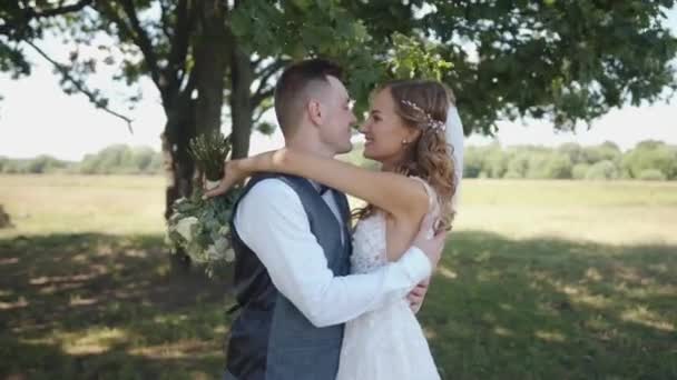 Noiva feliz e noivo com um suporte de buquê abaixo de uma árvore em um gramado verde e abraço. Movimento lento — Vídeo de Stock