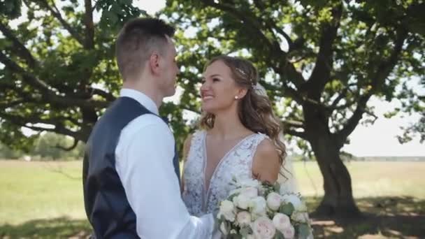 Boldog friss házasok állnak egy fa alatt, és a vőlegény orron csókolja a menyasszonyt egy csokorral a kezében egy gyönyörű csipke ruhában. Lassú mozgás. — Stock videók