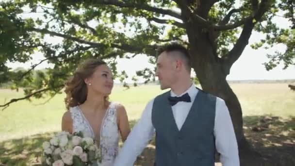 Happy bride and groom with a bouquet stand under a tree on a green lawn and hug. Slow motion — 图库视频影像