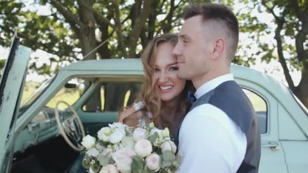 Bielorrússia. Região de Gomel. Agosto - 21, 2021: O noivo feliz abraça sua noiva rindo com um buquê enquanto está perto de um carro retro debaixo de uma árvore — Vídeo de Stock