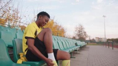 Belarus, Minsk - 20 Haziran 2021: Futbol formalı şirin siyah bir kız stadyumun tribününde oturur ve futbol çoraplarını giyer. Yavaş çekim