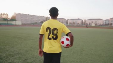 Futbol takımı üniformalı genç siyah bir kız elinde bir topla sahaya çıkıyor ve sırıtarak geriye bakıyor.