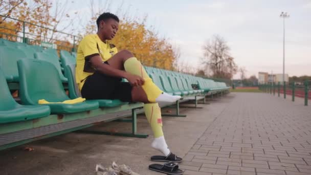 Vitryssland, Minsk - 20 juni 2021: Allvarlig ung svart flicka sitter på stadion åskådare tribun bär i sitt lag uniform och binda snören på hennes fotbollsskor — Stockvideo