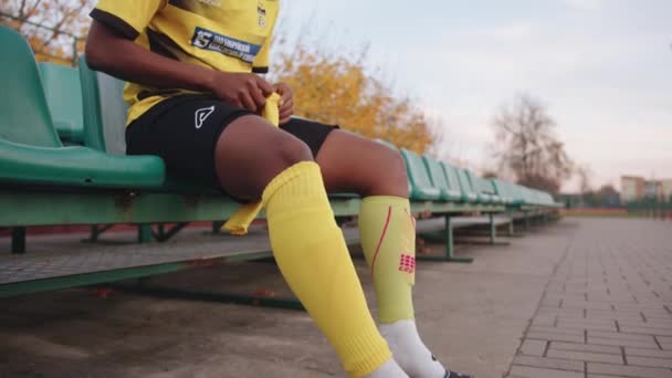 Vitryssland, Minsk - 20 juni 2021: En söt ung svart tjej i en fotbollströja sitter på åskådartribun i stadens stadion och sätter på sig fotbollsstrumpor. Långsamma rörelser — Stockvideo