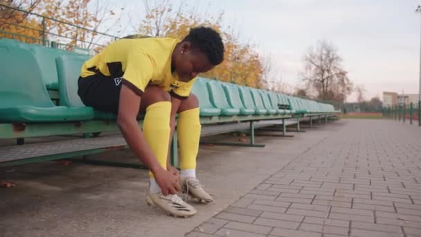Vitryssland, Minsk - 20 juni 2021: Allvarlig ung svart flicka sitter på stadion åskådare tribun bär i sitt lag uniform och binda snören på hennes fotbollsskor — Stockvideo