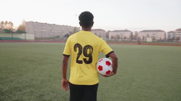 Una joven negra con uniforme de equipo de fútbol cruza el campo con una pelota en la mano y mira hacia atrás sonriendo — Vídeo de stock