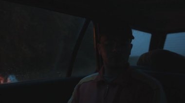 Gözlüklü ve şapkalı genç bir adam arabada oturur ve yağmur damlalarıyla ormanın yakınındaki şenlik ateşine bakar.