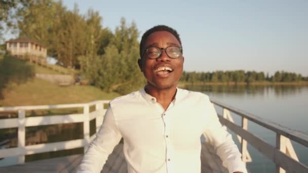 Ένας νεαρός μαύρος στέκεται στην προβλήτα δίπλα στο ποτάμι και κοιτάζει τον ουρανό με τα χέρια του απλωμένα. Έννοια ελευθερίας. Αργή κίνηση — Αρχείο Βίντεο