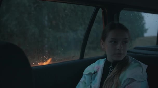 Скромна молода дівчина сидить в машині біля багаття в дощовий день і дає інтерв'ю в мікрофон з ґудзиком — стокове відео