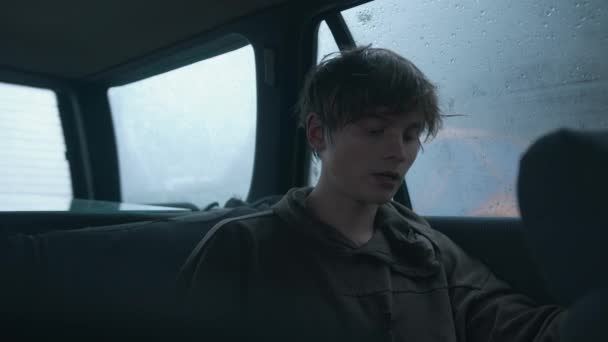 Hipster-buscando joven se sienta en un coche bajo la lluvia y da una entrevista en el micrófono lavalier conectado a la ropa — Vídeo de stock