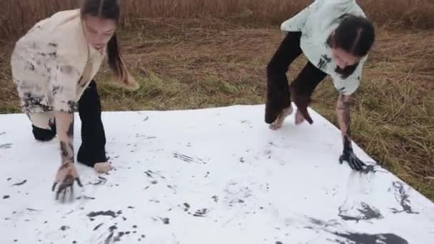 Zwei junge Mädchen führen in einem Weizenfeld einen alternativen Tanz auf, wobei ihre Hände mit schwarzer Farbe beschmiert sind und Spuren auf Kleidung und Leinwand hinterlassen — Stockvideo