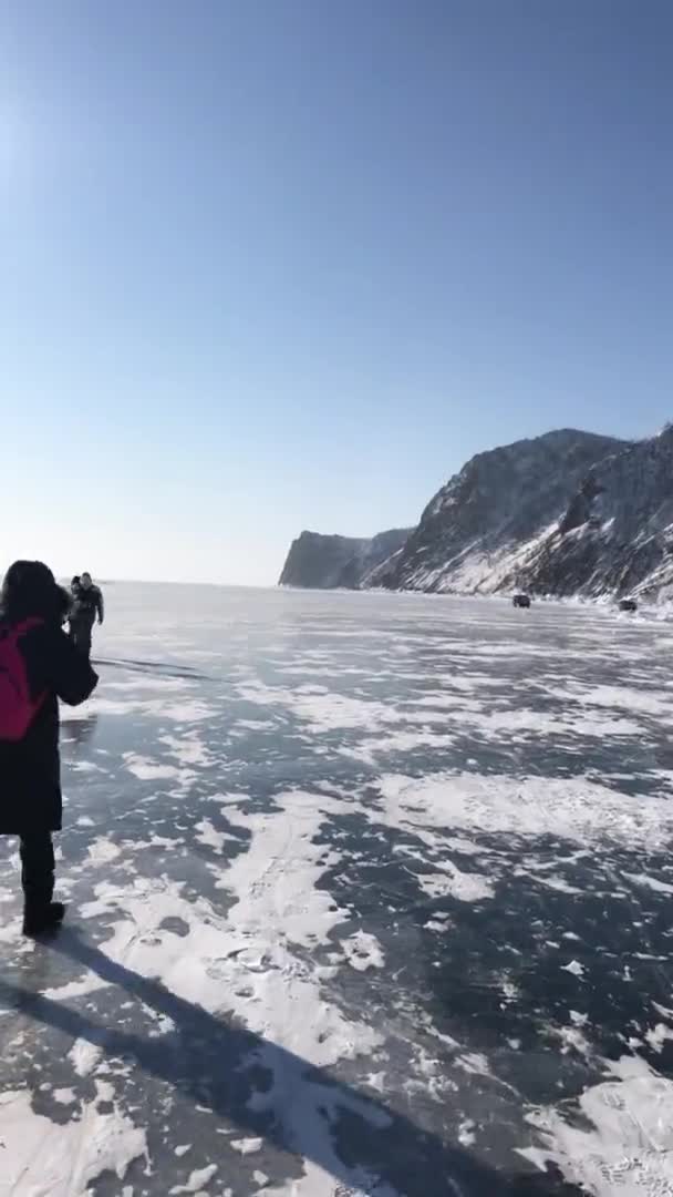 Un grupo de turistas va sobre el hielo transparente del lago Baikal y el panorama de la costa rocosa contra el cielo azul soleado. Vídeo vertical — Vídeos de Stock