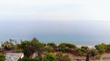 Sahildeki ağaçların arasındaki çatıların havadan görünüşü ve güneşli bir günde mavi denizde yalnız beyaz bir yat.