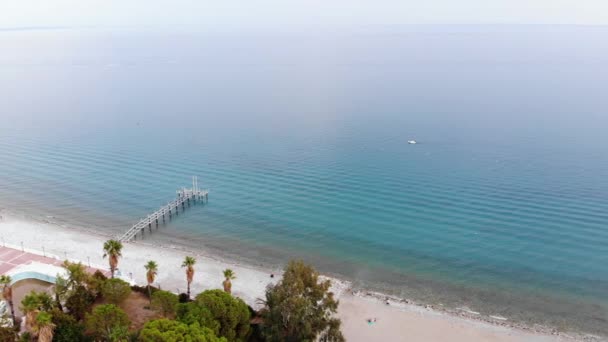 Luchtfoto van wit strand met pier en palmbomen in recreatiegebied en eenzaam wit jacht tussen de rustige blauwe golven van helder blauwe zee — Stockvideo