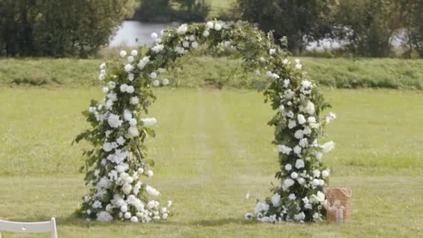 Весільна арка з ніжними білими квітами і гілочками зелені, що змішуються з вітру на газоні біля річки — стокове відео