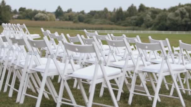 Σειρές από λευκές καρέκλες σε ένα καταπράσινο γκαζόν περιτριγυρισμένες από δέντρα για τους καλεσμένους σε μια γαμήλια τελετή — Αρχείο Βίντεο