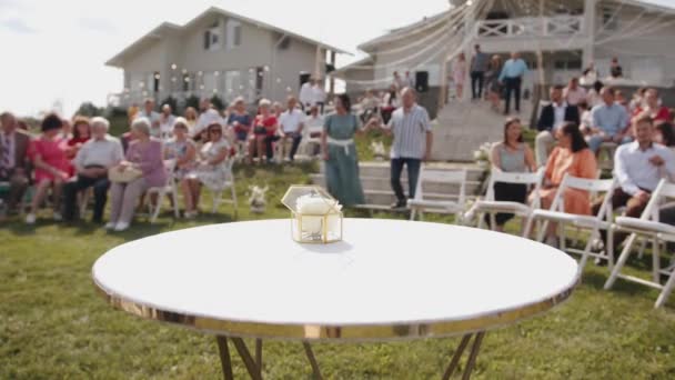Una vista de una caja de vidrio con anillos en la mesa y fondo borroso con invitados en sillas en el césped por una casa de campo antes de la boda — Vídeos de Stock