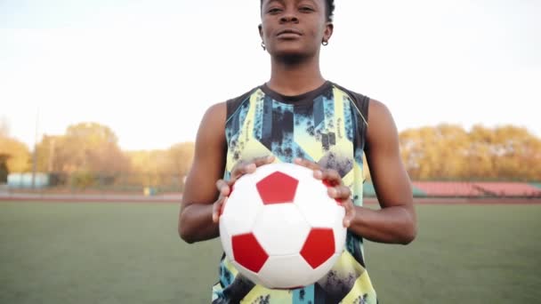 Een jonge Afro-Amerikaanse vrouw in een mouwloos T-shirt staat op een voetbalveld met een bal in haar handen en toont haar hand gebald in een vuist — Stockvideo