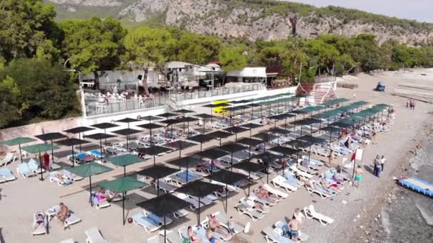 Aerial view of rows of sun loungers with sunbathing tourists in a recreation area on a mountain coast and people swimming in the sea — Stockvideo