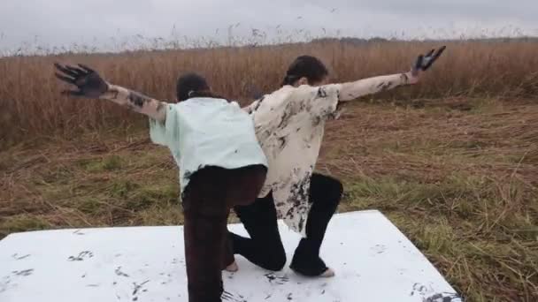 Две молодые девушки танцуют альтернативный танец на пшеничном поле, руки мажут черной краской и оставляют следы на одежде и холсте. — стоковое видео