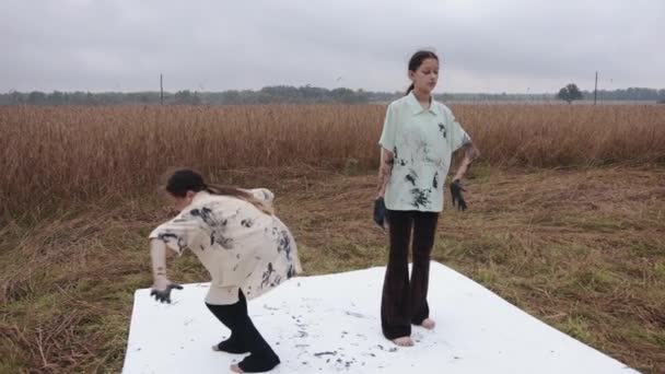 Дві молоді дівчата виконують альтернативний танець на пшеничному полі своїми руками, замазаними чорною фарбою і залишають сліди на одязі і полотні — стокове відео