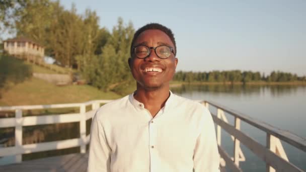 Młody czarny facet w okularach stoi na drewnianym pomoście nad brzegiem rzeki i zamykając oczy cieszy się słonecznym letnim dniem. Zwolniony ruch — Wideo stockowe