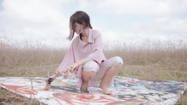 Beautiful young girl stained with paint sits on his haunches on a large canvas in the field and paints an abstract picture — Stockvideo