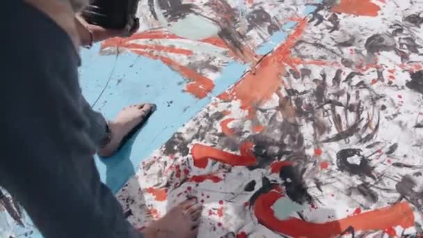 A young girl draws an abstract picture on a large canvas in the field and outlines black paint over her bare foot on top of the drawing — Video Stock