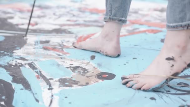 A young artist goes with bare feet stained with paint on a large canvas lying on the field and draws an abstract picture — 비디오