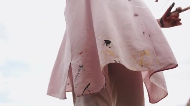 Een jong meisje staat in een veld en haar roze wijde shirt bevlekt met verf fladdert in de wind. Kunst en creativiteit concept. Langzame beweging — Stockvideo