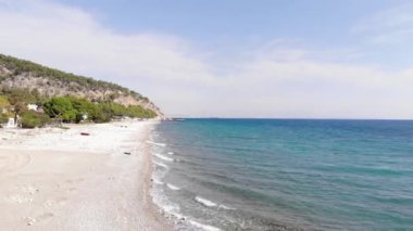 Ağaçların arasında binalar ve beyaz sahilde dalgalanan dalgalarla birlikte açık mavi deniz manzarası.