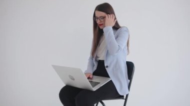 Gözlüklü, kendine güvenen genç iş kadını bir sandalyede otururken dizüstü bilgisayarda çalışıyor ve saçını düzleştiriyor. Yavaş çekim