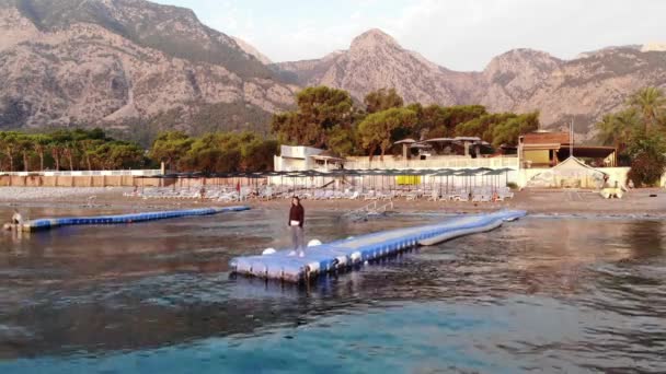 Aerial view of a pensive young girl standing on the plastic floating pier in the sea against the background of a mountain town on the coast. Retreat concept. solitude in nature — Stockvideo