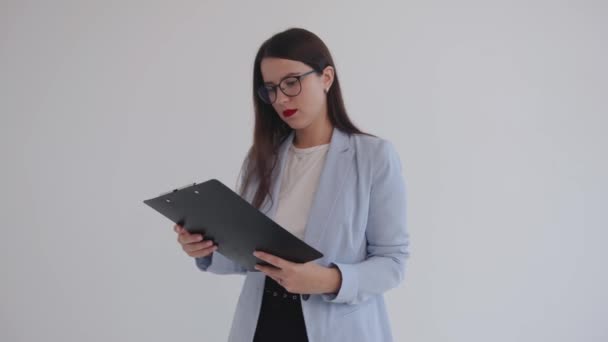 Beautiful business woman in black glasses examines documents pinned on a clipboard and purses her lips in displeasure — стоковое видео